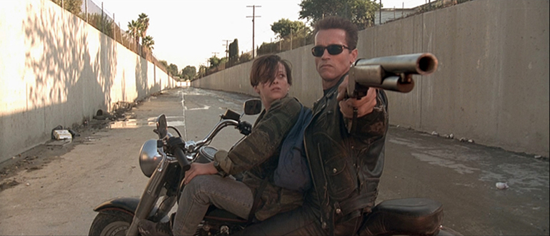 Arnold as the terminator in Terminator 2: Judgment Day aims his shotgun back at the crashed semi driven by the T1000