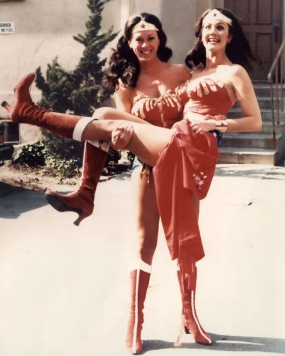 Jeannie Epper and Lynda Carter are dressed in matching Wonder Woman costumes while Jeannie carries Lynda bride style. Both looks super happy and Lynda looks surprised and delighted