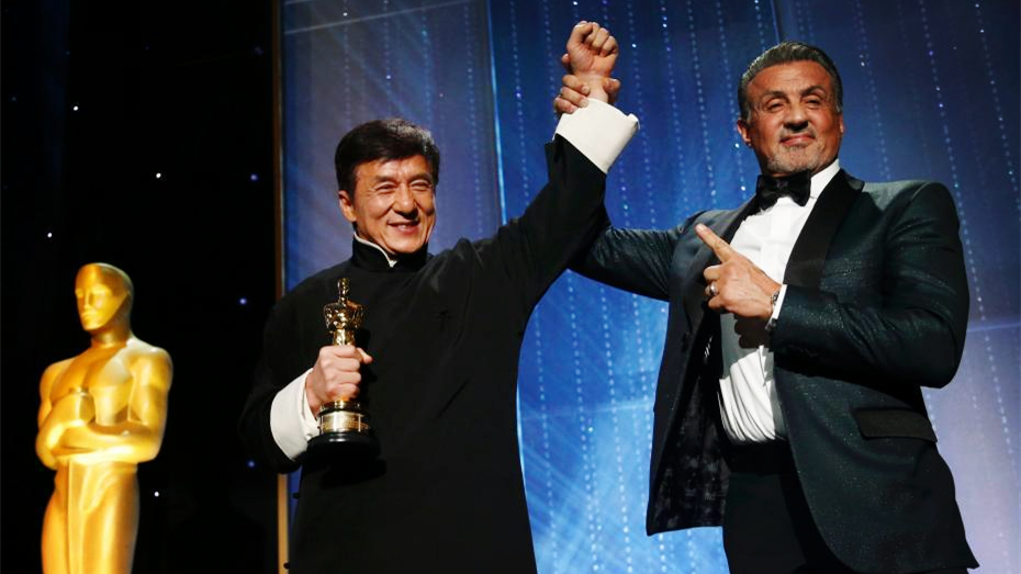 Sylvester Stallone points at Jackie Chan holding his Oscar, as Stallone raises Jackie's arm like a champ